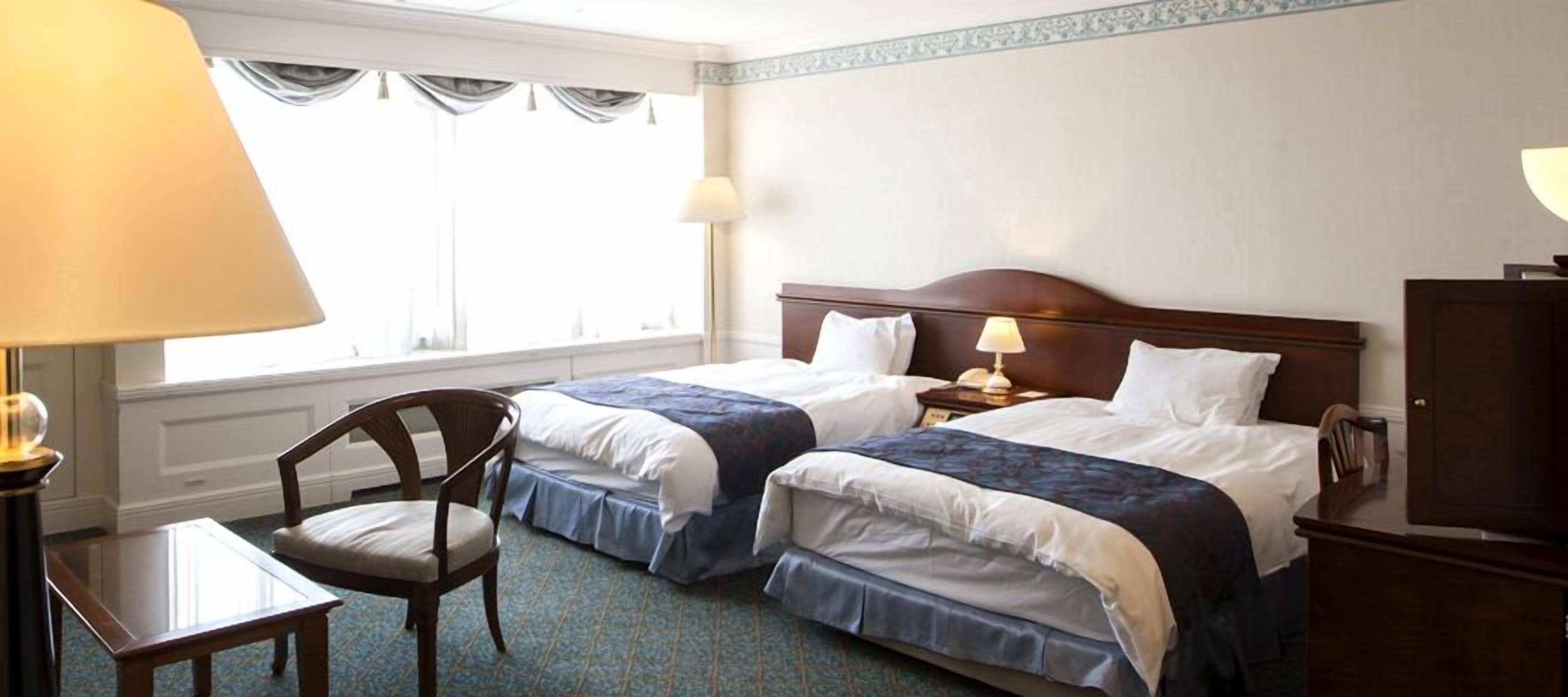 A hotel room with two beds, a desk, a chair, and a lamp.