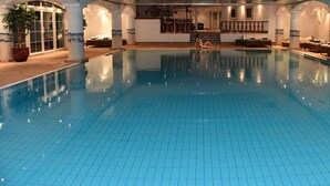 A large indoor swimming pool with tiled flooring, lounge areas with seating, and a staircase leading to an upper level. - Grand Hôtel du Golf & Palace (Crans-Montana)