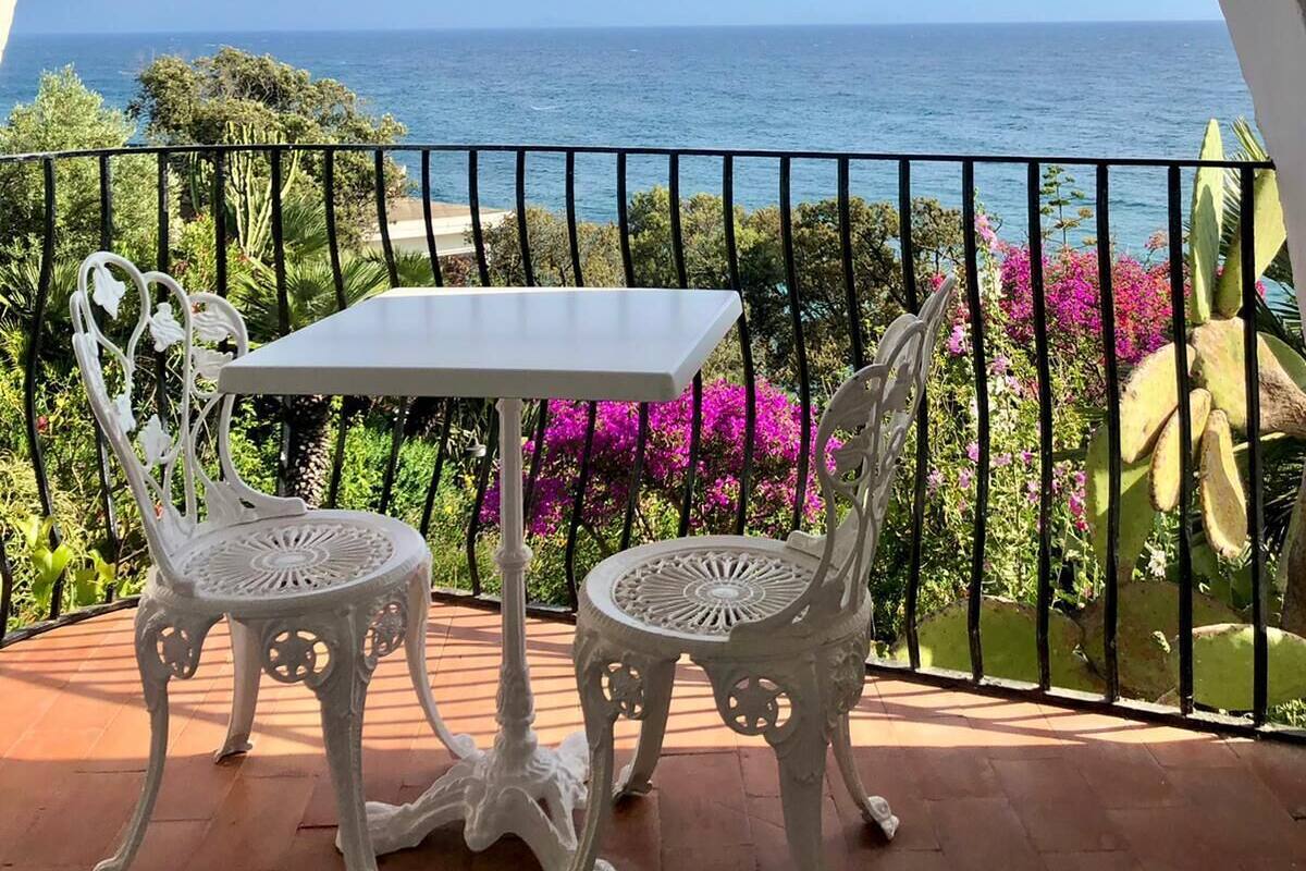 A balcony with a white table and two ornate chairs, overlooking a coastal view with the sea and greenery.