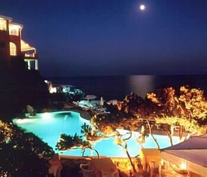 Een nachtzicht van een resort met een zwembad, ligbedden en een gebouw met verlichte ramen.