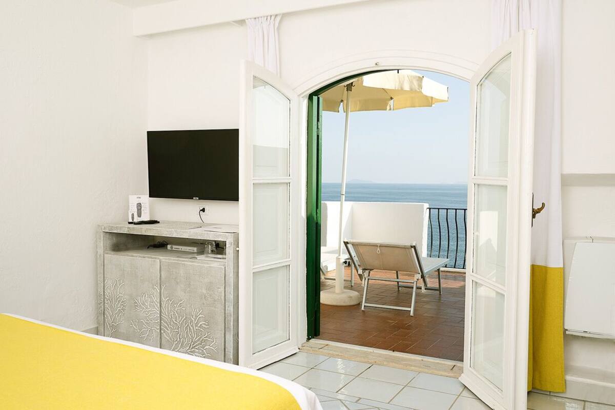 A bedroom with a bed, a TV, a desk, and a balcony with a chair and umbrella.