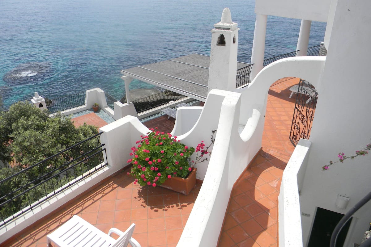 A balcony with a view of the sea, a white chair, and a planter with pink flowers.