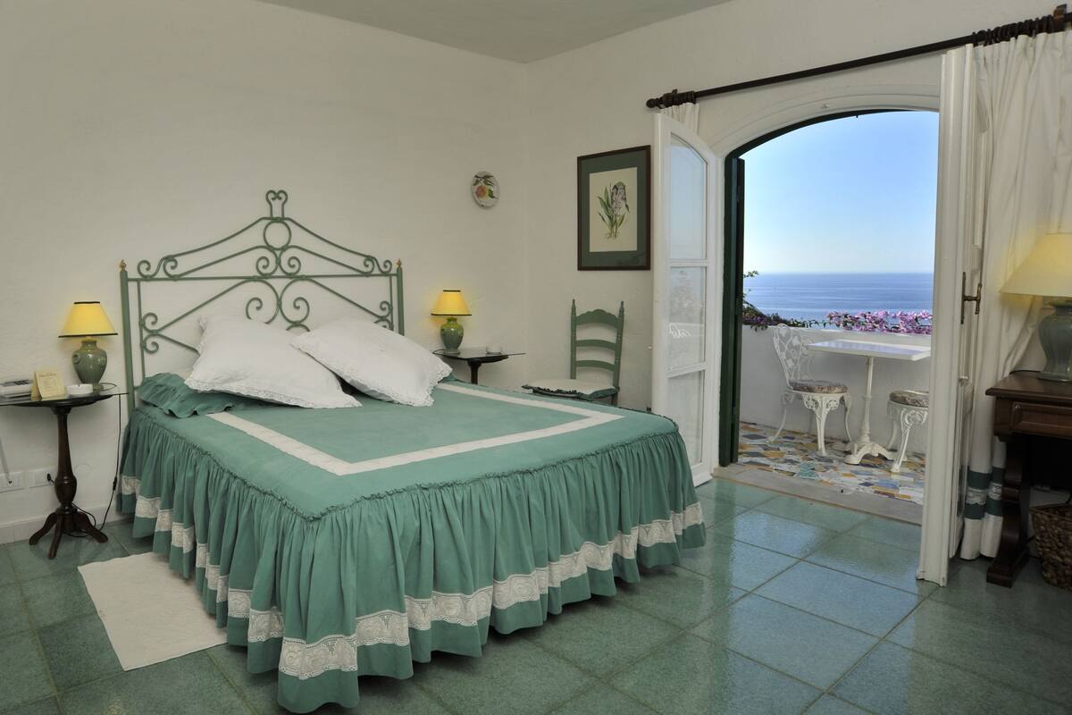 A bedroom with a bed, two bedside tables, a chair, and a view of the sea through an open door.