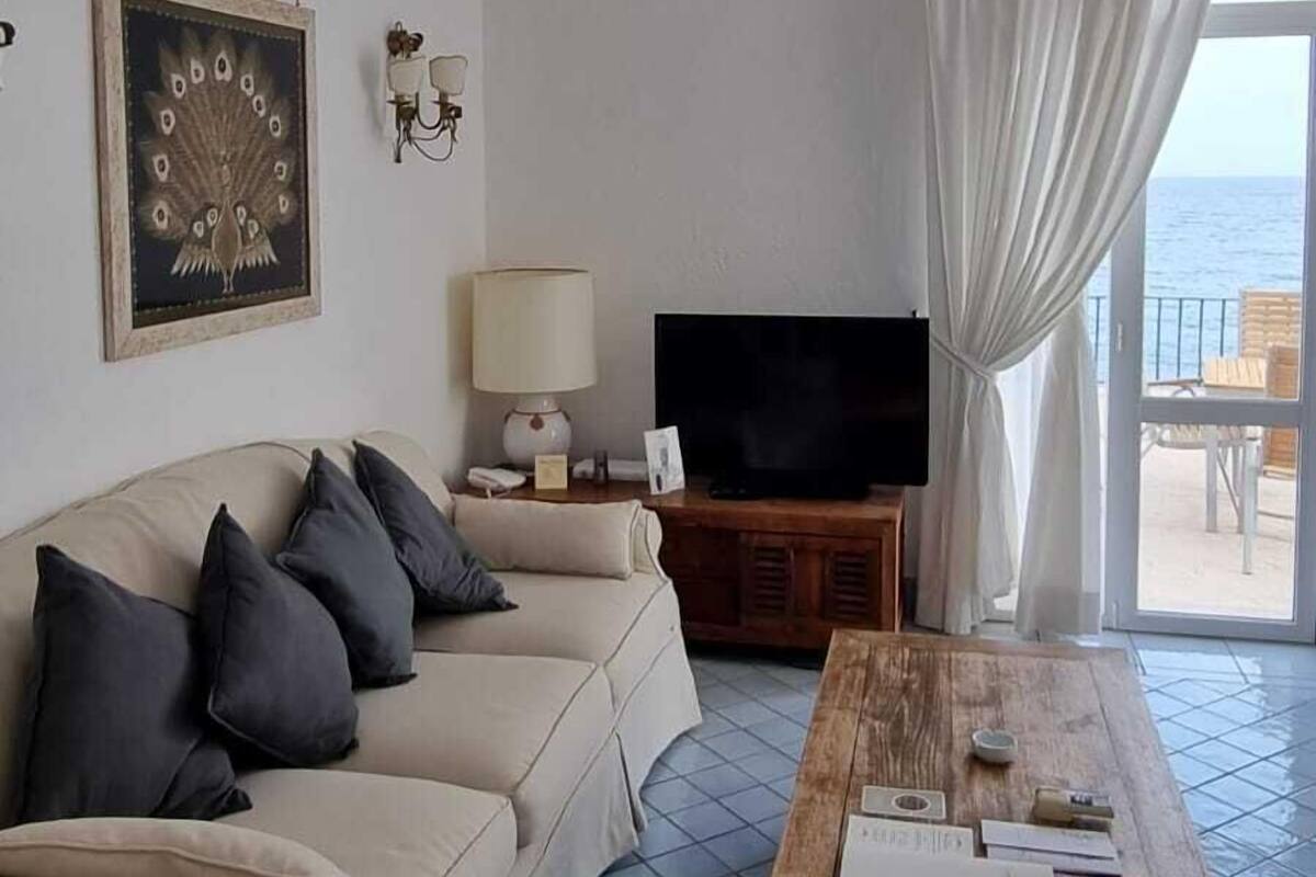 A living room with a beige sofa, a wooden coffee table, a flat-screen TV, and a view of the sea through a window with white curtains.
