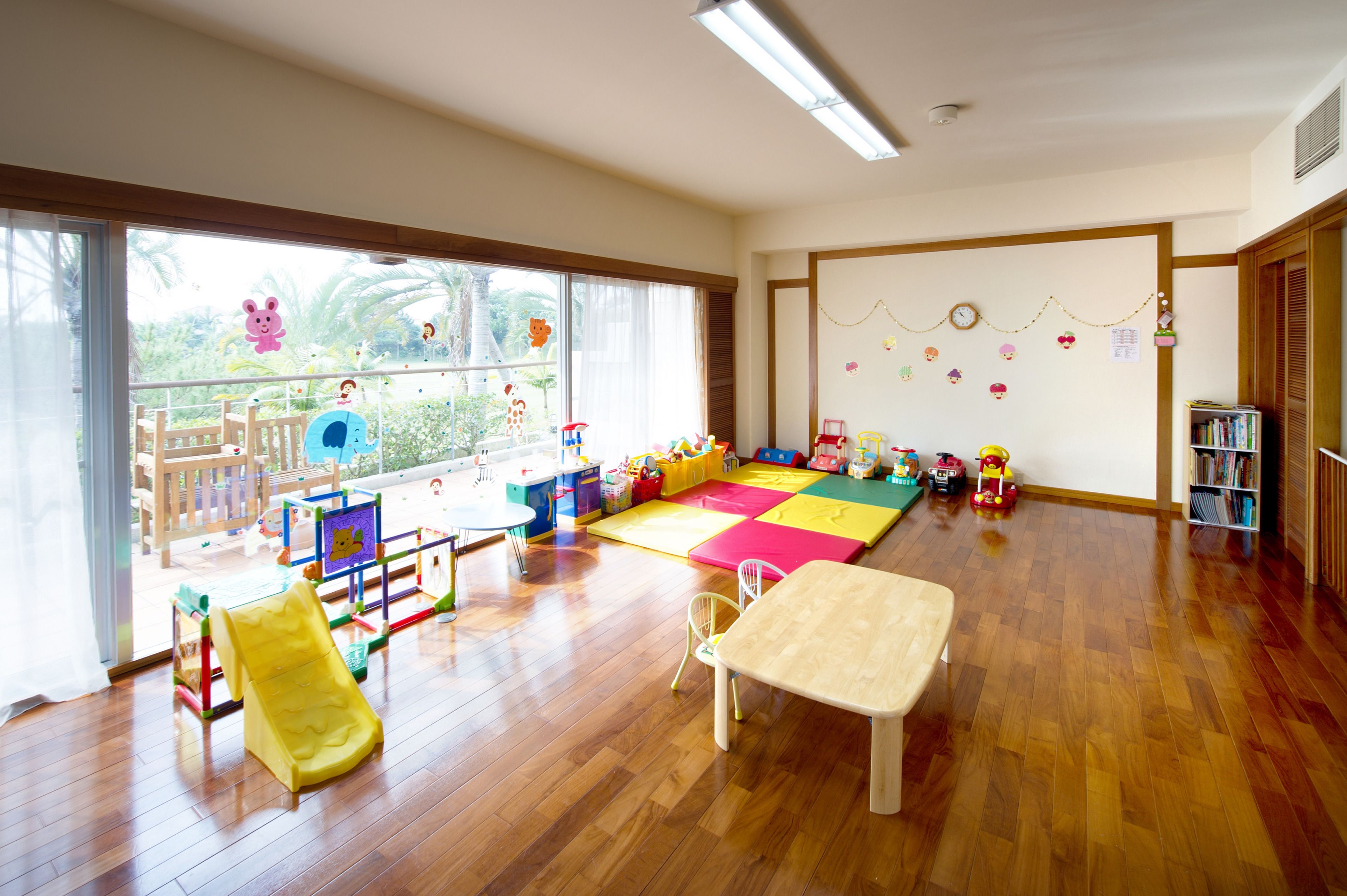 A spacious room with a large window, wooden flooring, a play area with colorful mats and toys, and a bookshelf.