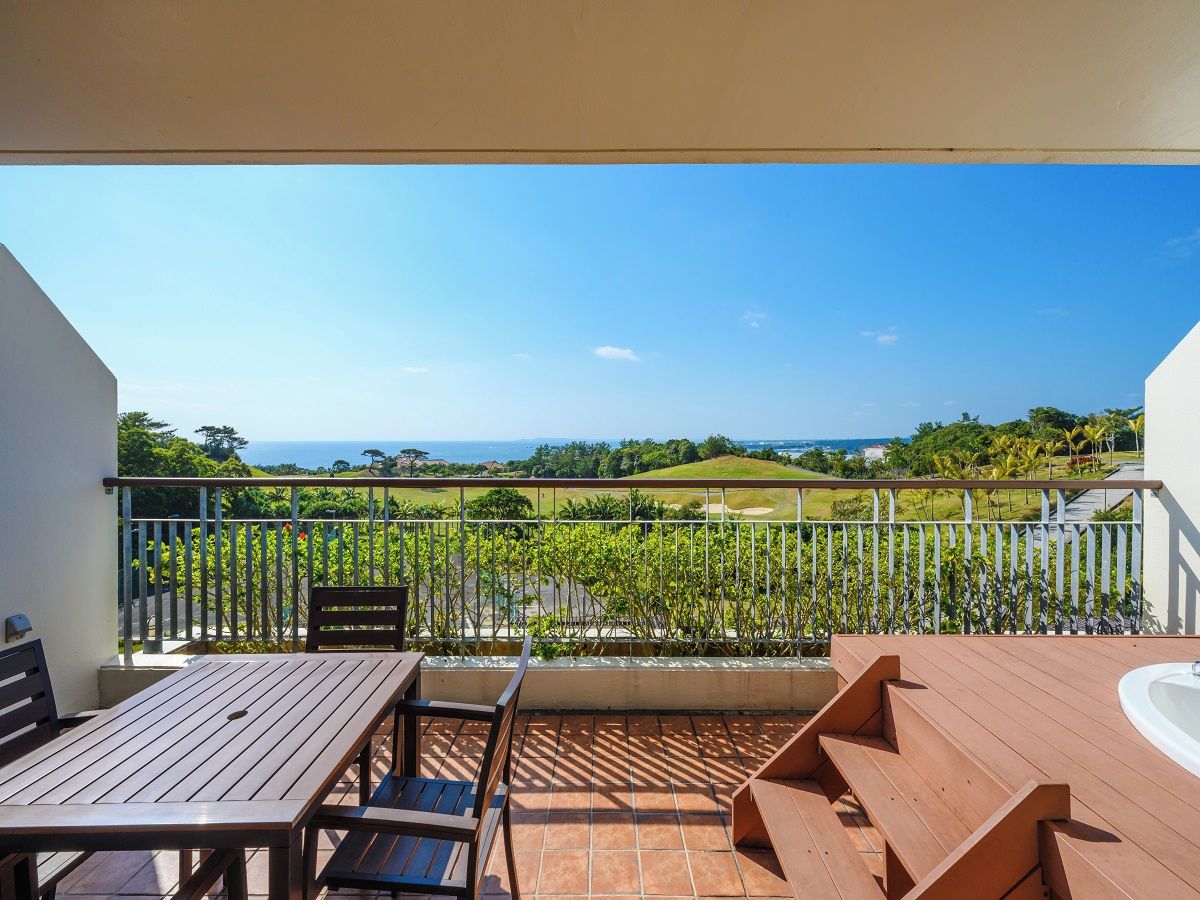 A balcony with a wooden table and chairs, a white bathtub, and a view of a golf course and ocean.