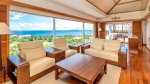 A spacious living room with large windows offering a view of the ocean. There are two sofas and a coffee table. - Kanucha Bay Hotels & Villas (Nago)