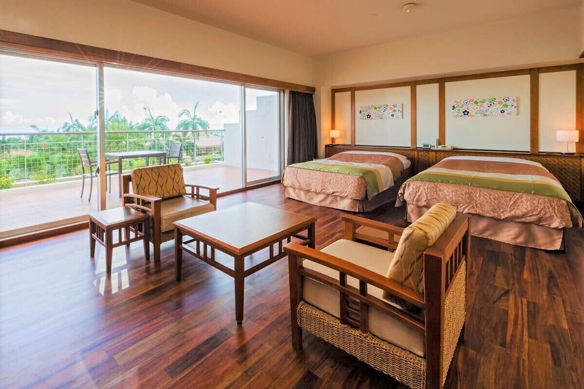 A hotel room with two beds, a balcony with a table and chairs, and a view of palm trees.