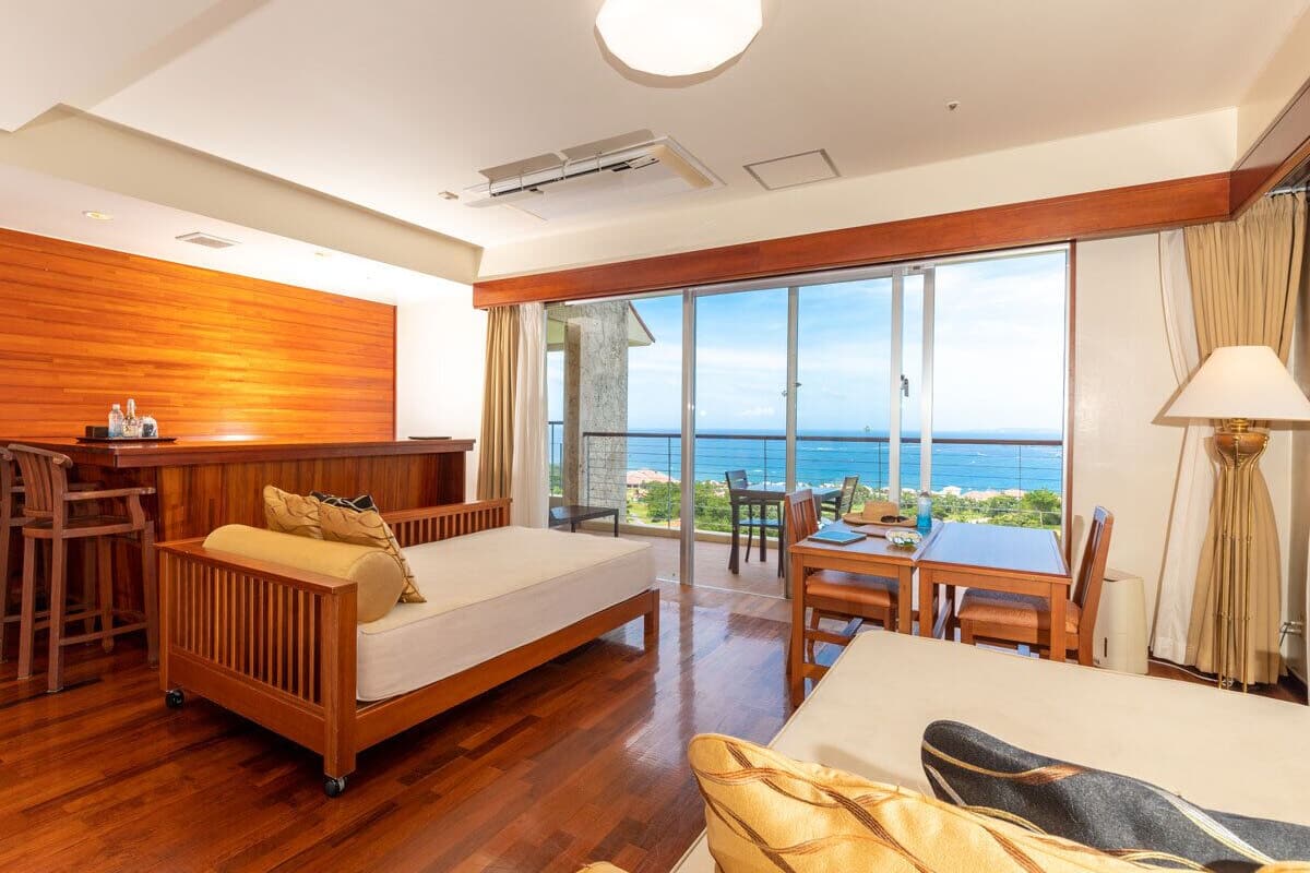 A hotel room with a wooden floor, a bed, a dining table, and a view of the ocean.