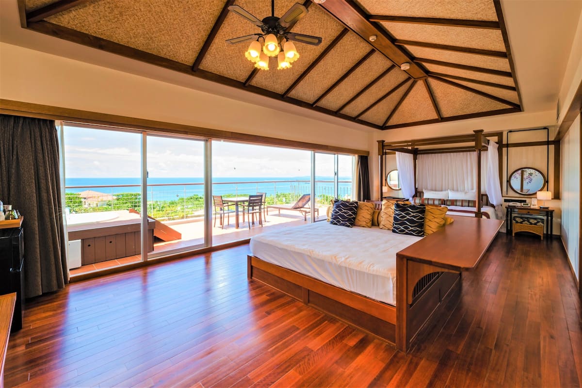 A spacious bedroom with a large bed, a ceiling fan, and a balcony with a view of the sea.