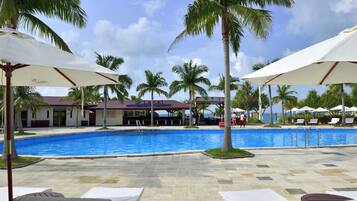 A pool area with lounge chairs and umbrellas, surrounded by palm trees and a building with a red roof.