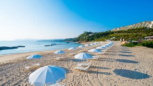A beach with rows of white lounge chairs and umbrellas, a clear sky, and a building with balconies in the background. - Kanucha Bay Hotels & Villas (Nago)