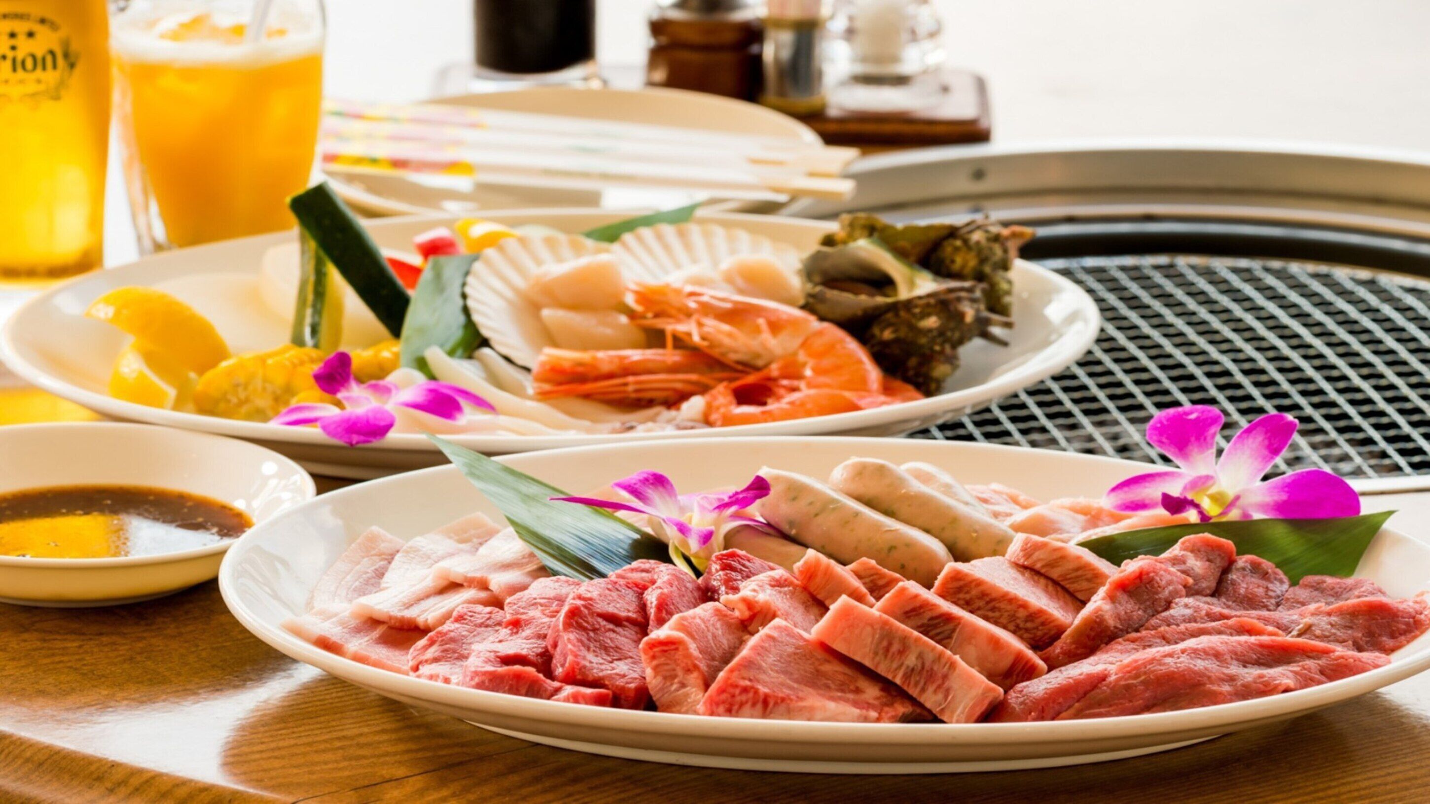 A table with plates of raw meat, seafood, and a bowl of sauce, along with a glass of beer and a plate of sliced fruit.