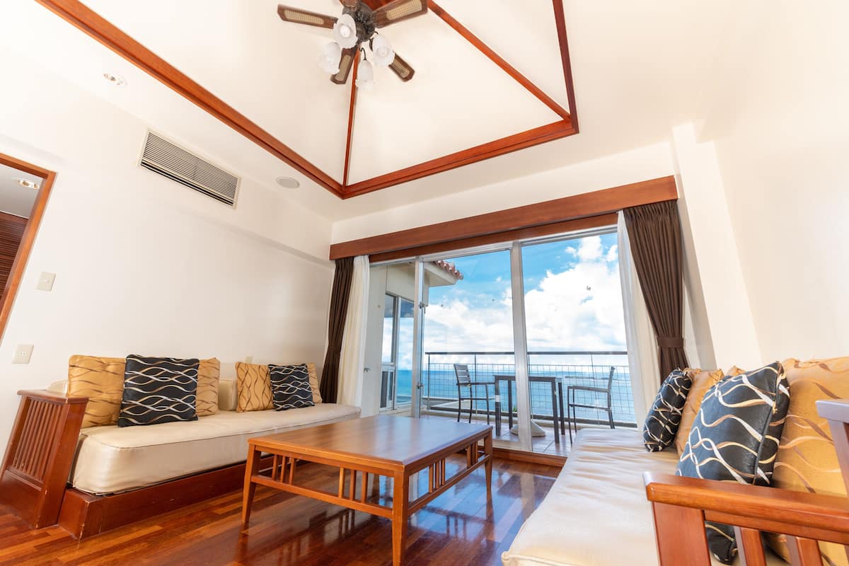 A spacious living area with a wooden floor, a sofa with patterned cushions, a wooden coffee table, and a large window offering a view of the sea.