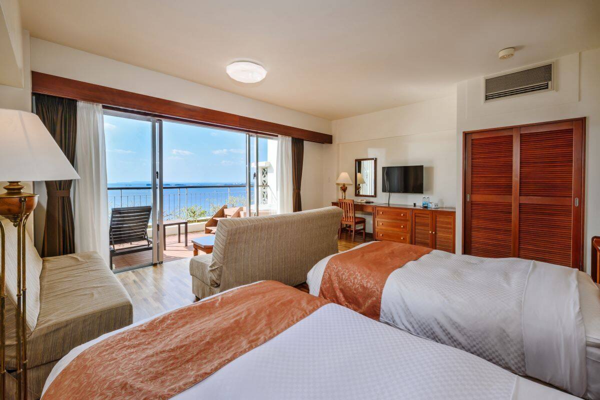 A hotel room with two beds, a desk, a chair, and a balcony with a view of the sea.