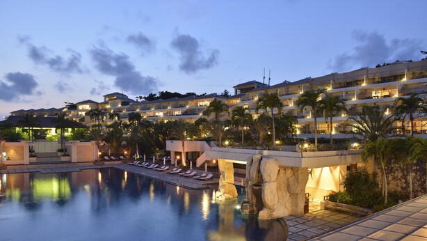 A large swimming pool with lounge chairs, palm trees, and a hotel building in the background. - Kanucha Bay Hotels & Villas (Nago)