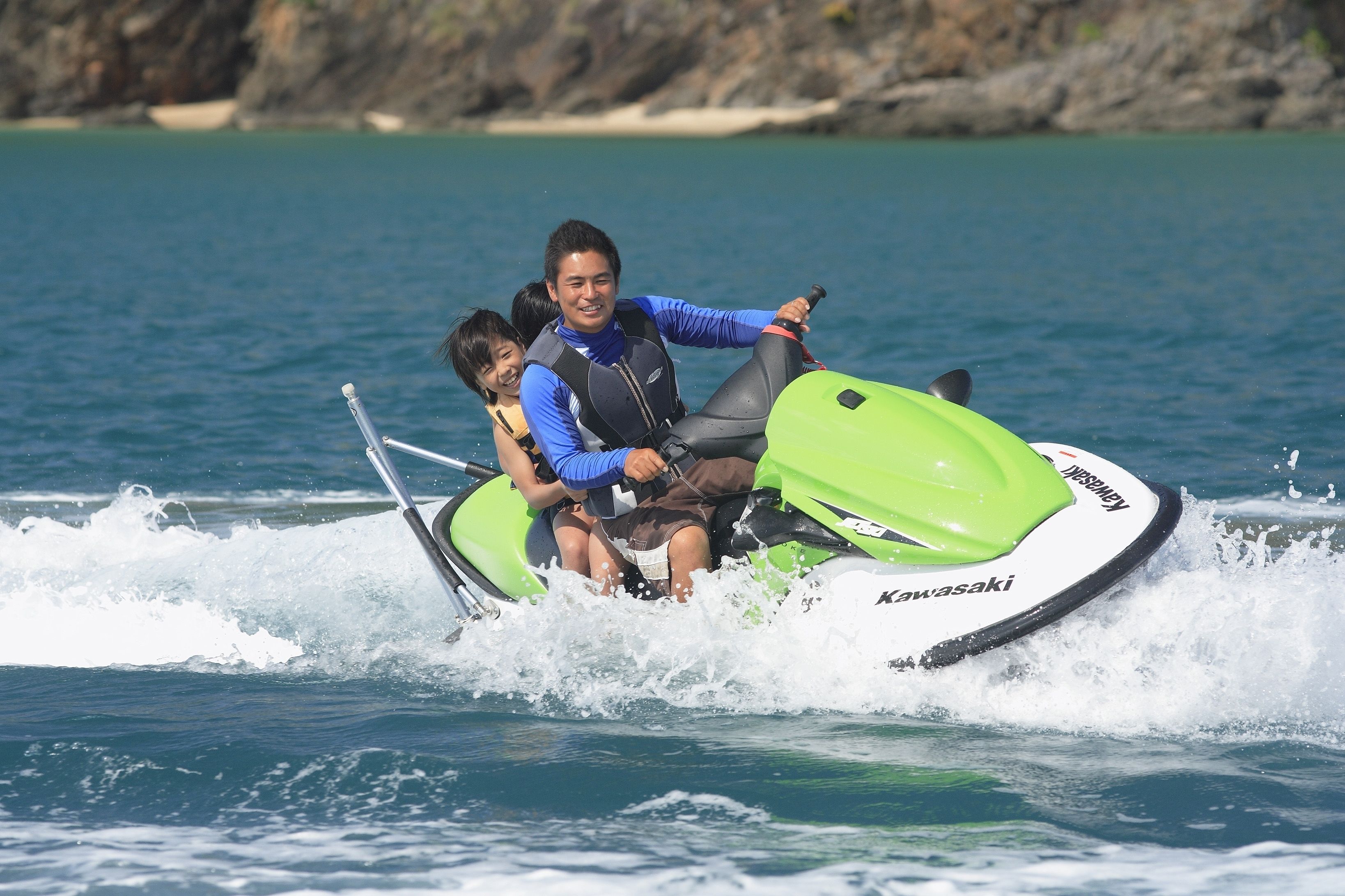 A person riding a green and white Kawasaki jet ski on a body of water with a rocky shoreline in the background.