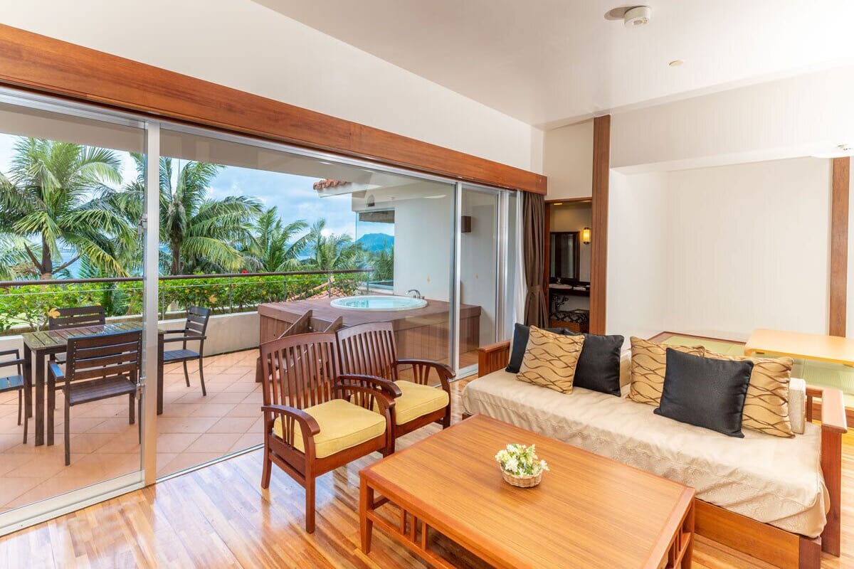 A modern living room with a sofa, wooden chairs, a coffee table, and a sliding glass door leading to a balcony with a pool and palm trees.