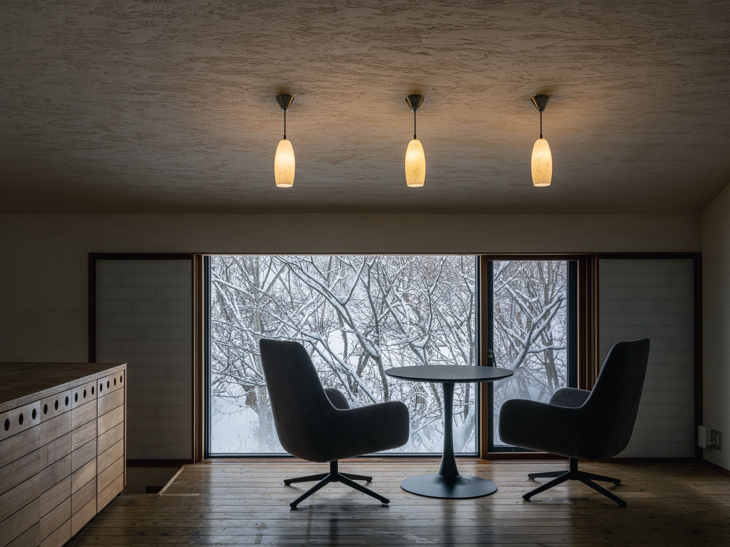 Quarto moderno com uma janela grande, duas cadeiras pretas, uma mesa redonda e luminárias pendentes.
