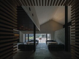 A modern interior with a sloped ceiling, wooden beams, and a stone floor. There are seating areas with tables and benches.