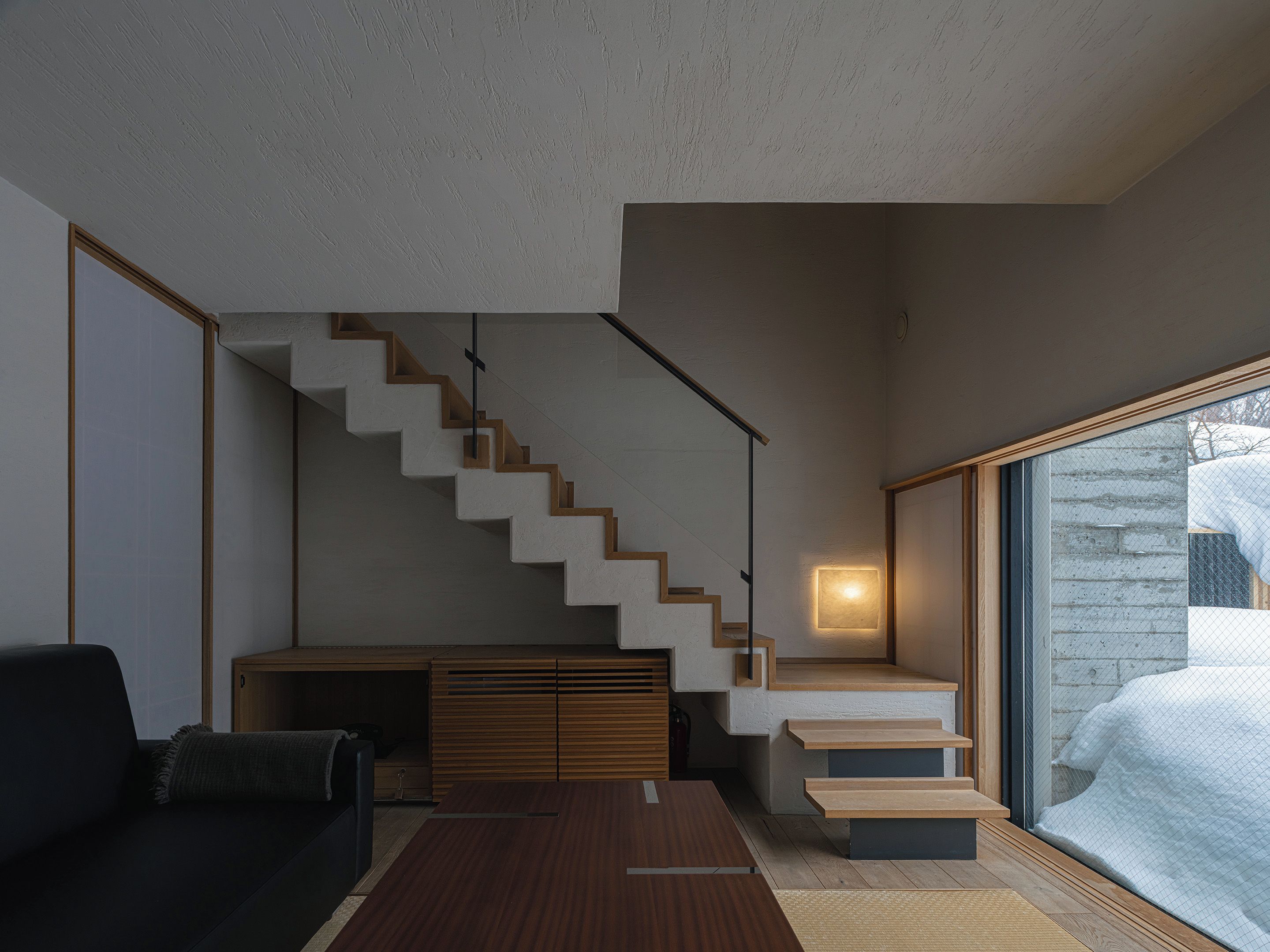 A modern living room with a staircase, a sofa, a coffee table, and a large window with a view of a snowy landscape.