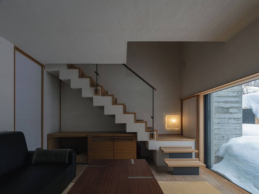 A modern living room with a staircase, a sofa, a coffee table, and a large window with a view of a snowy landscape.