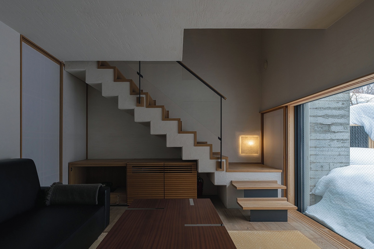 A modern living room with a staircase, a sofa, a coffee table, and a large window with a view of a snowy landscape.