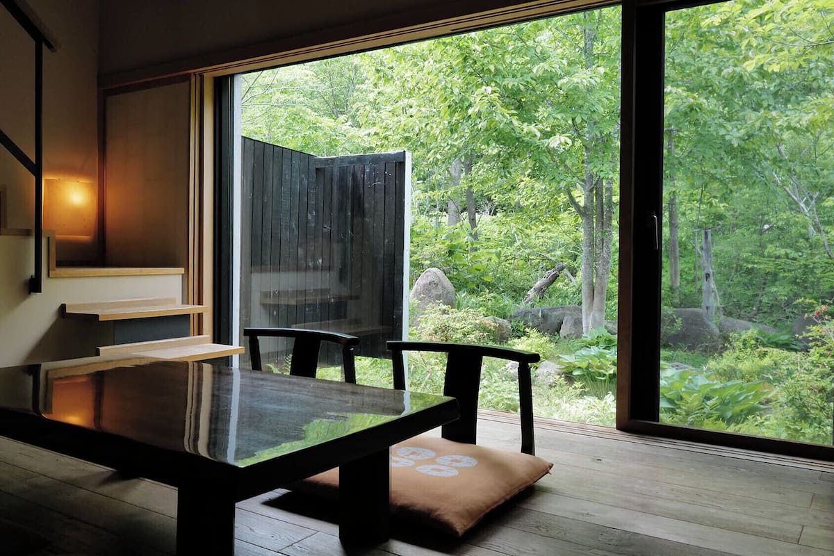 Un interior moderno con una mesa de madera, sillas, una escalera y un ventanal con vistas a un bosque.