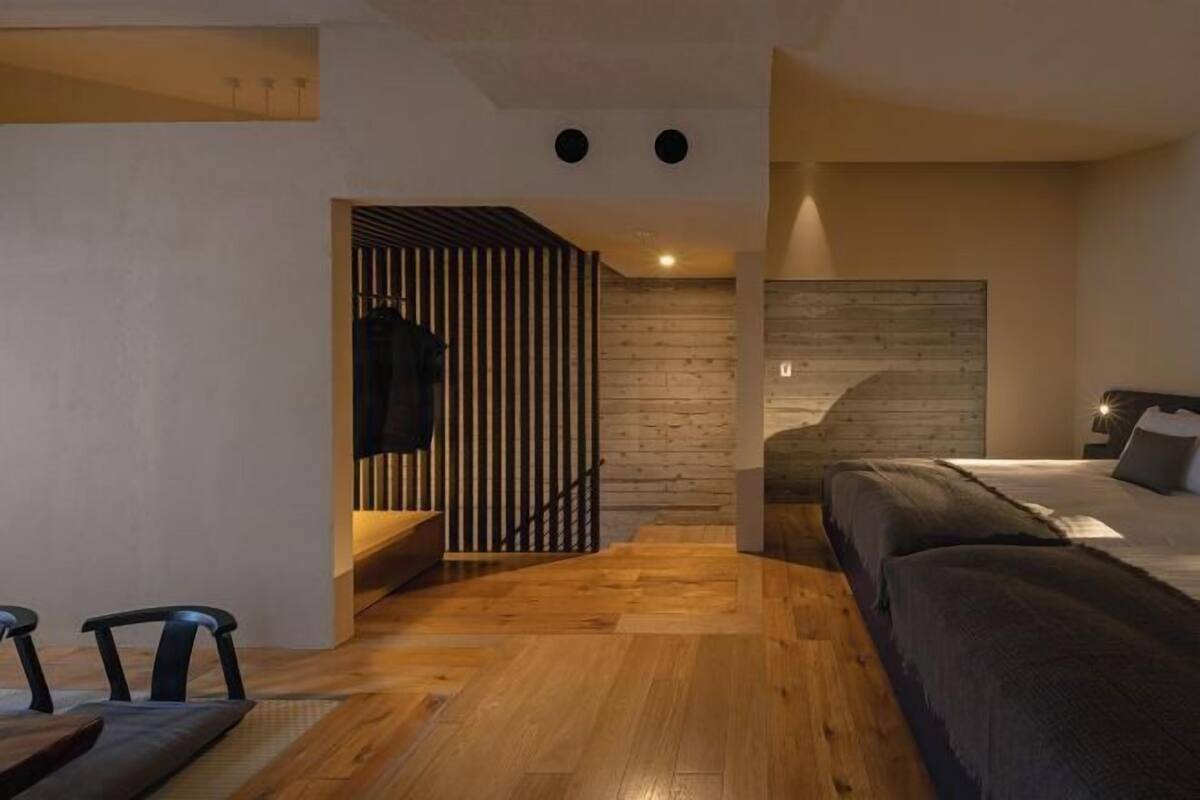 Un dormitorio moderno con suelo de madera, una cama con ropa de cama oscura, un televisor montado en la pared y una escalera que lleva a otra habitación.