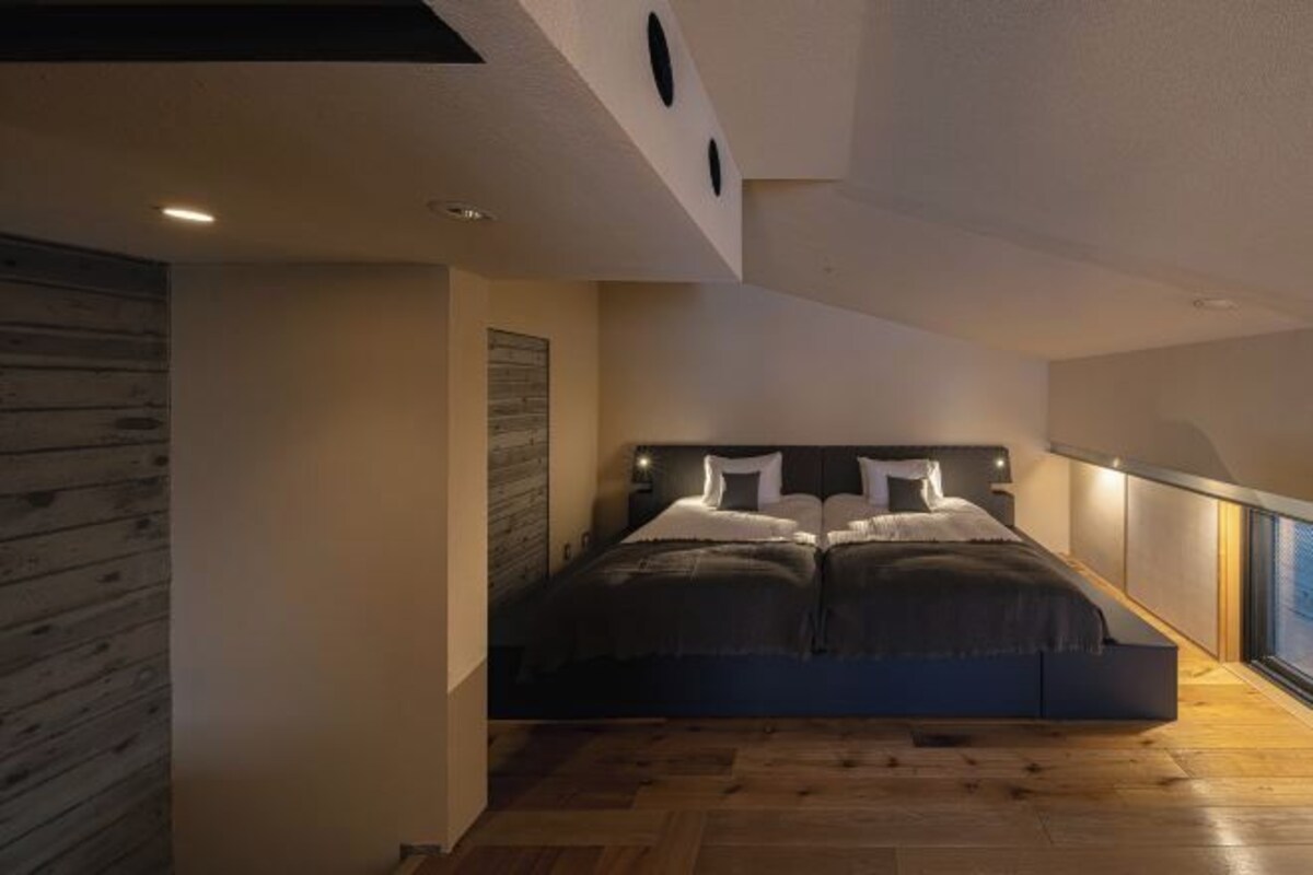 A bedroom with a large bed, wooden flooring, and a view through a window.