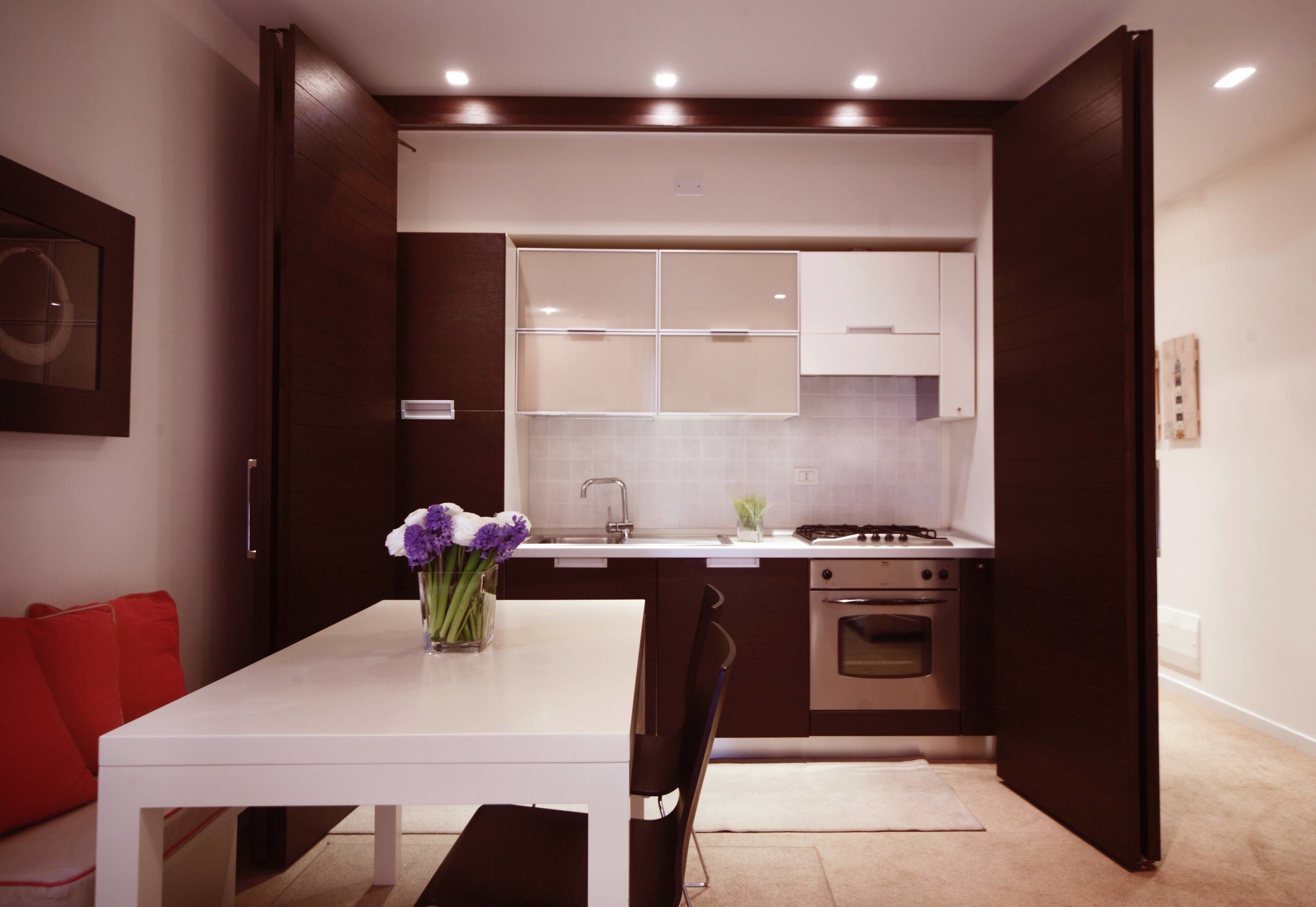 Una cocina moderna con armarios oscuros, encimera blanca y zona de comedor con mesa blanca y sillas rojas.