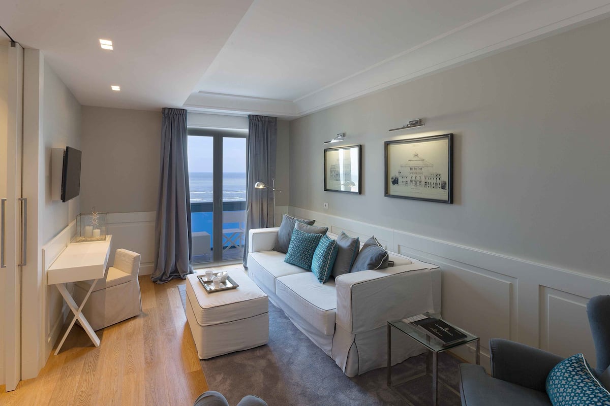 A modern hotel room with a balcony, a white sofa with patterned cushions, a small table, and a TV mounted on the wall.
