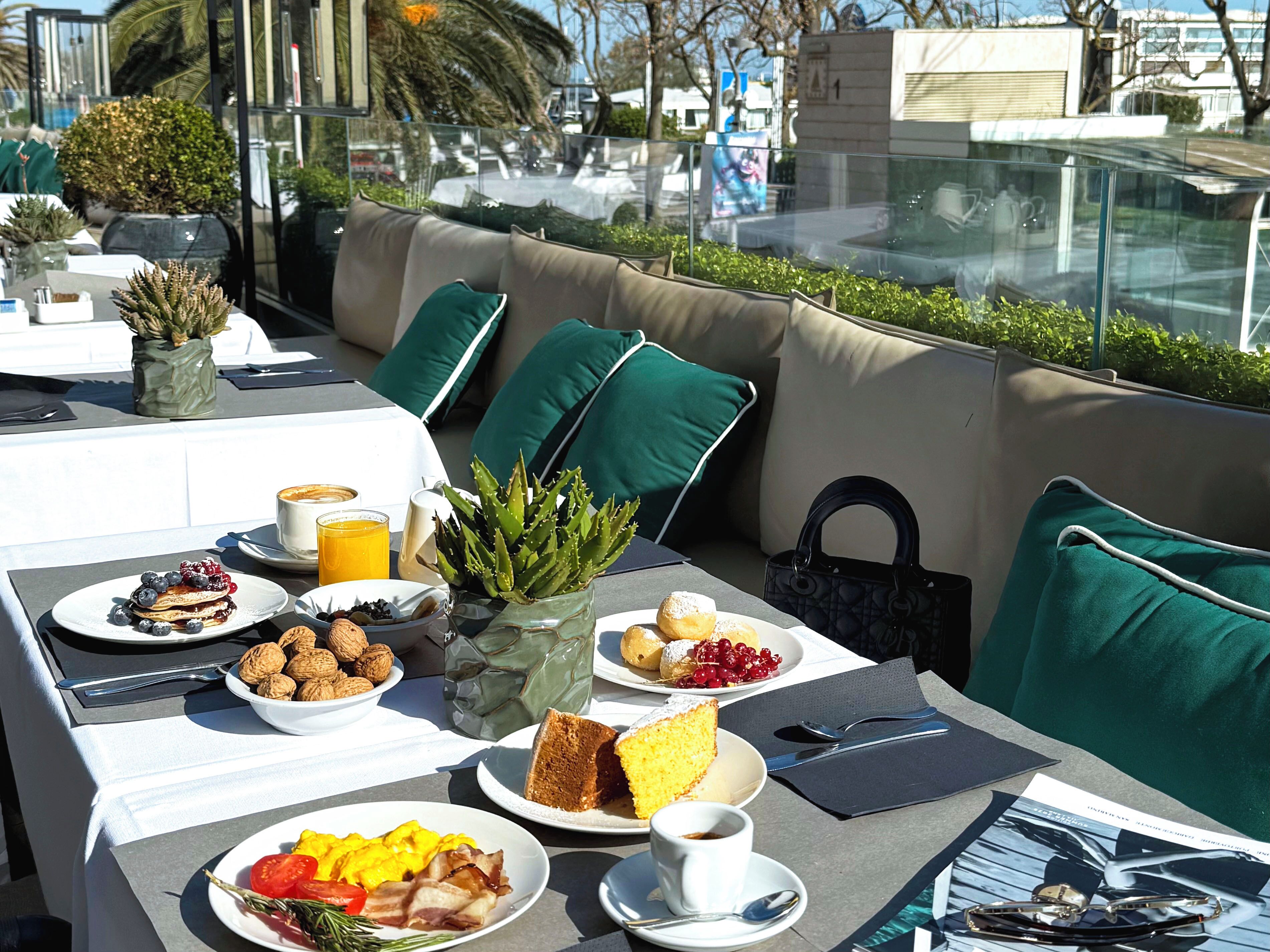 A table set with breakfast items, a green plant, and a handbag on a sunlit outdoor seating area.