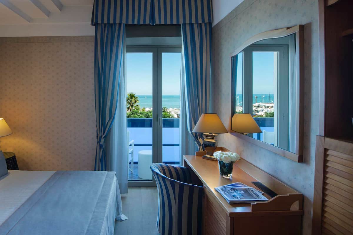 A hotel room with a bed, two bedside lamps, a desk with a chair, a window with a view of the sea, and a framed picture on the wall.