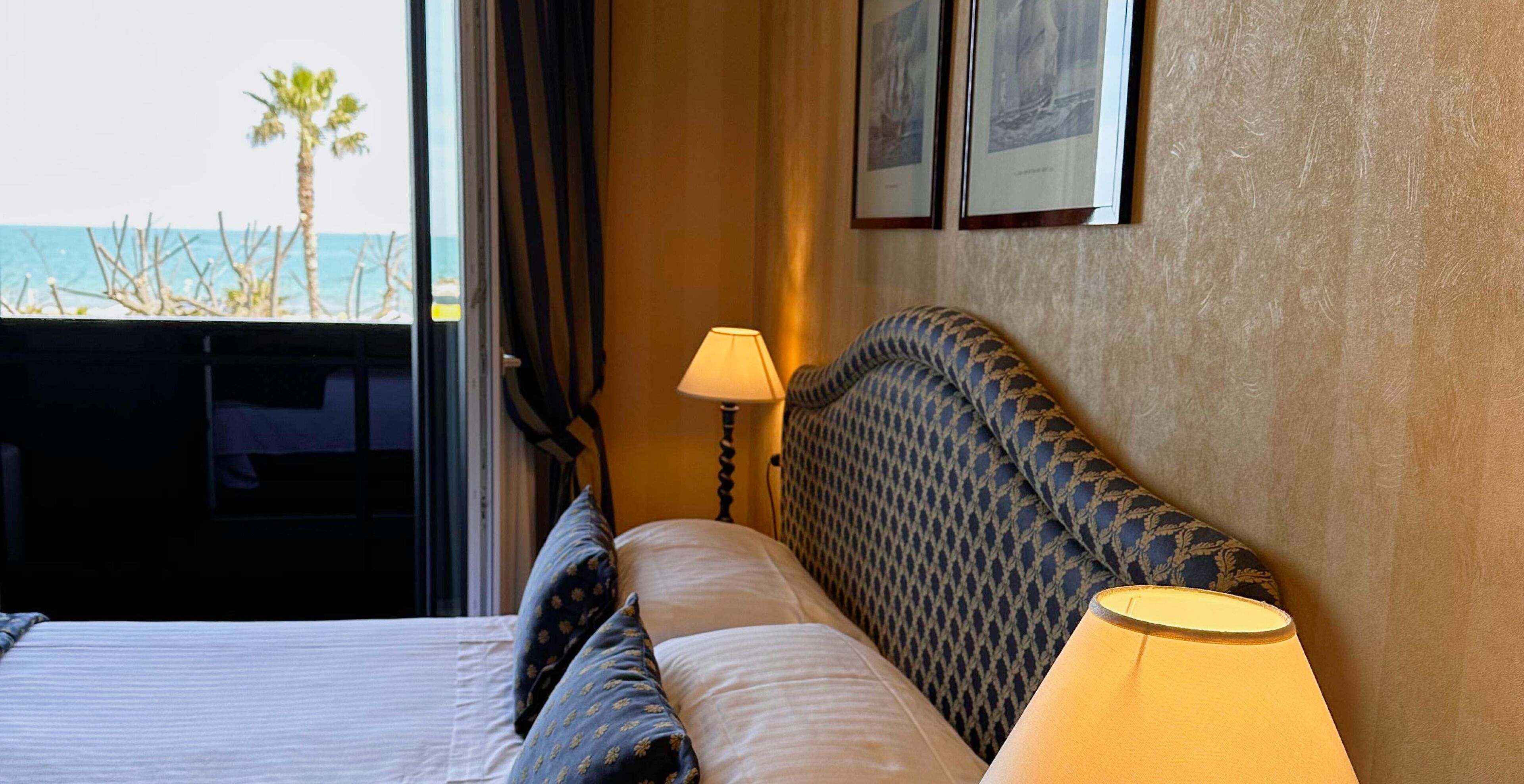 A hotel room with a bed, a nightstand with a lamp, a window with a view of the beach, and a wall with framed pictures.