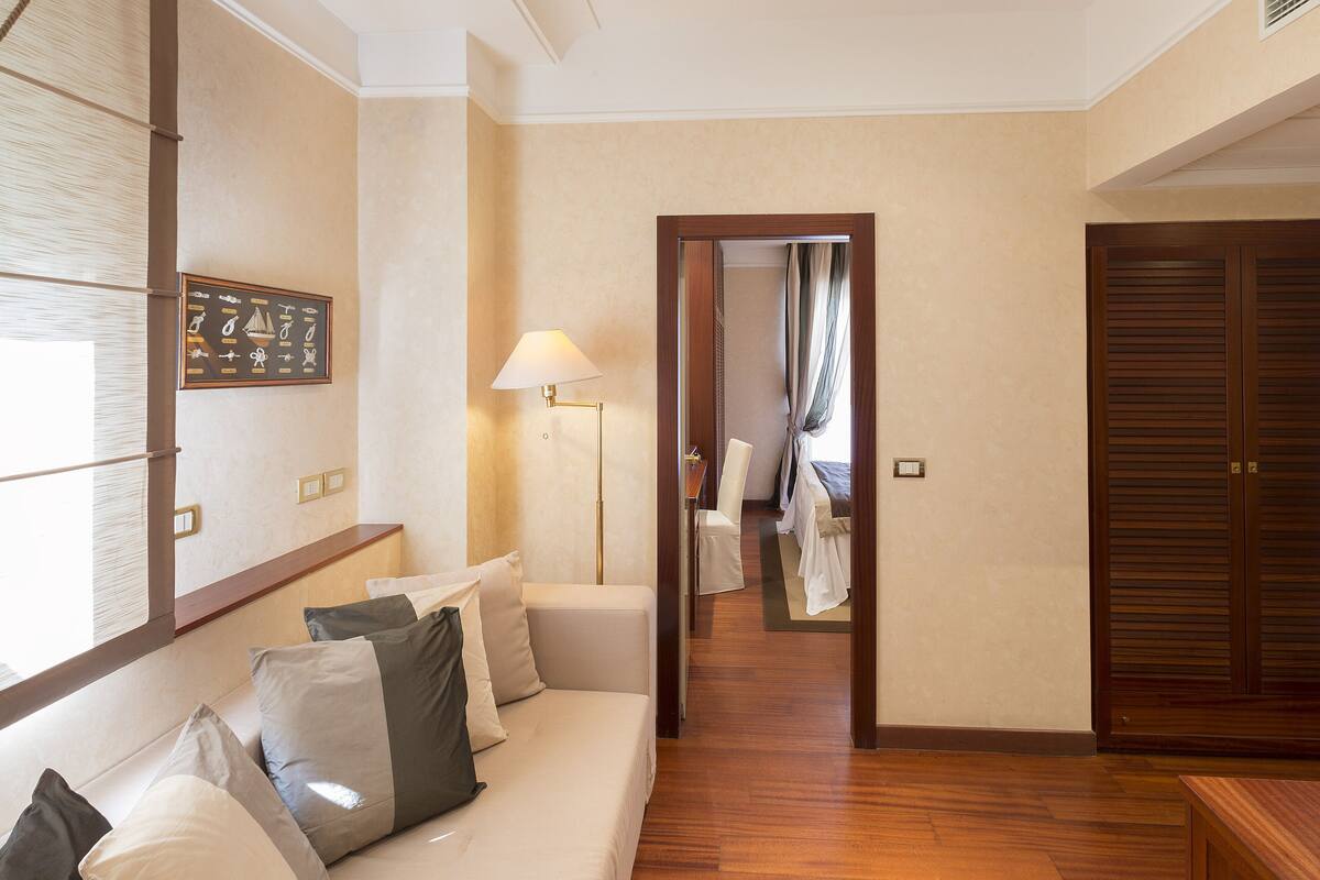 A hotel room with a wooden floor, a sofa with cushions, a desk, a lamp, and a wardrobe.