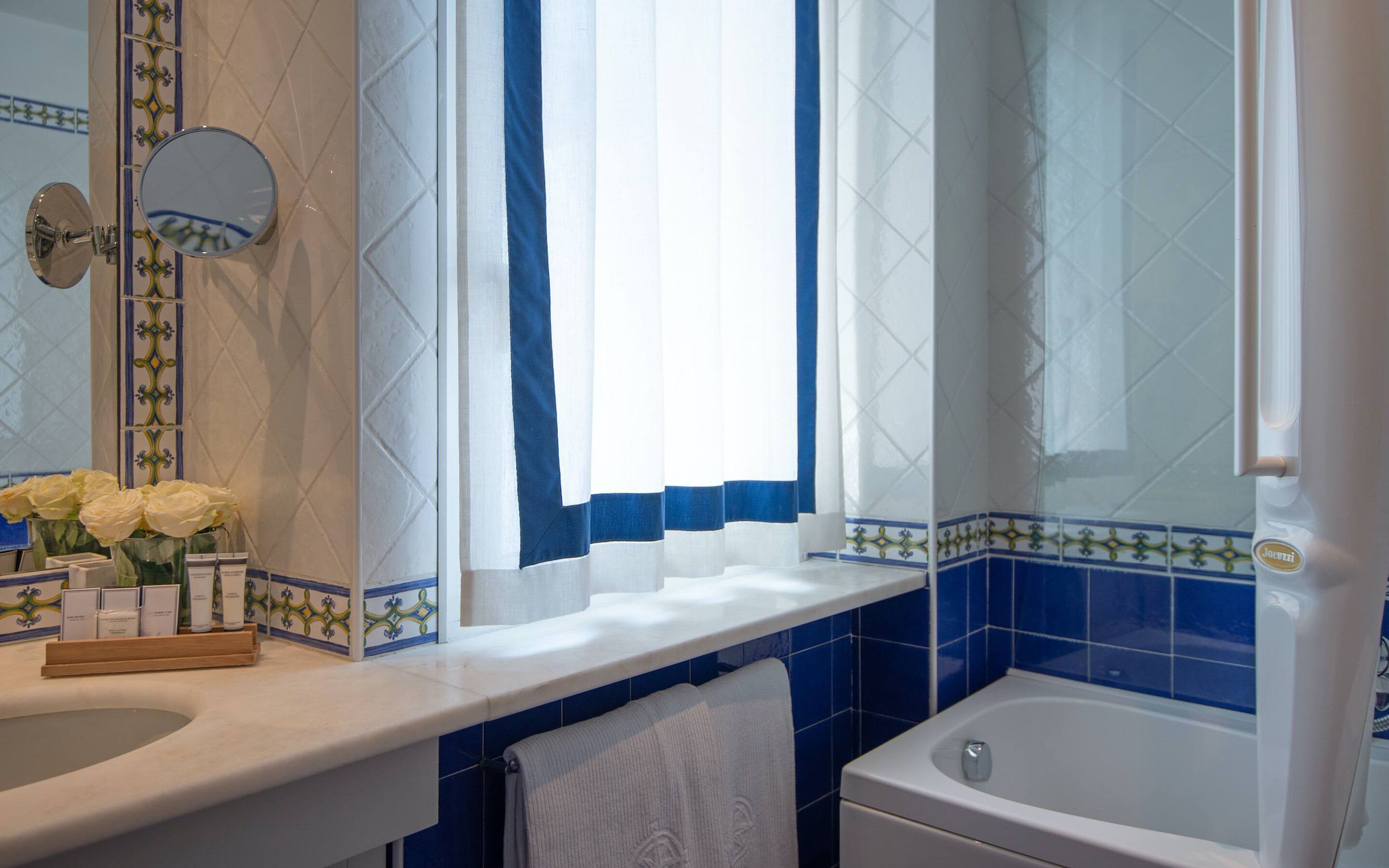 A bathroom with a sink, mirror, window with curtains, and a bathtub.