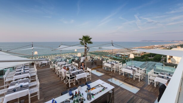 Un área de comedor al aire libre con mesas y sillas, con vista al mar y al paisaje urbano.