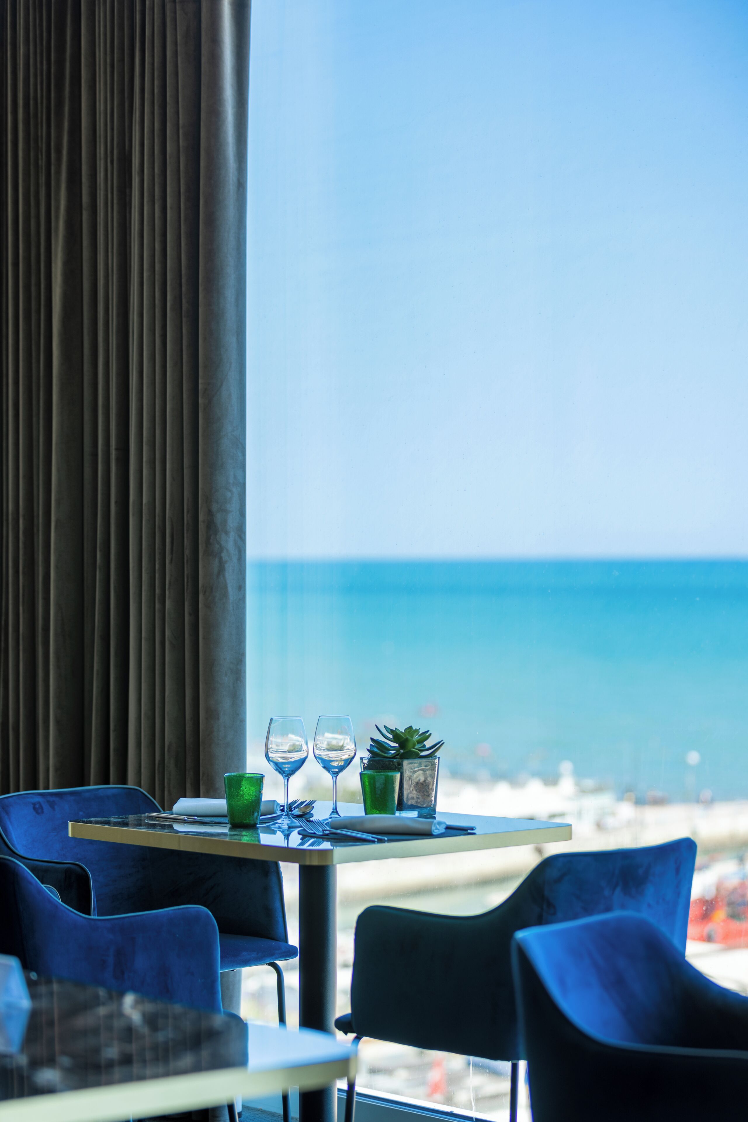 A dining area with a table set for two, a view of the sea, and blue chairs.