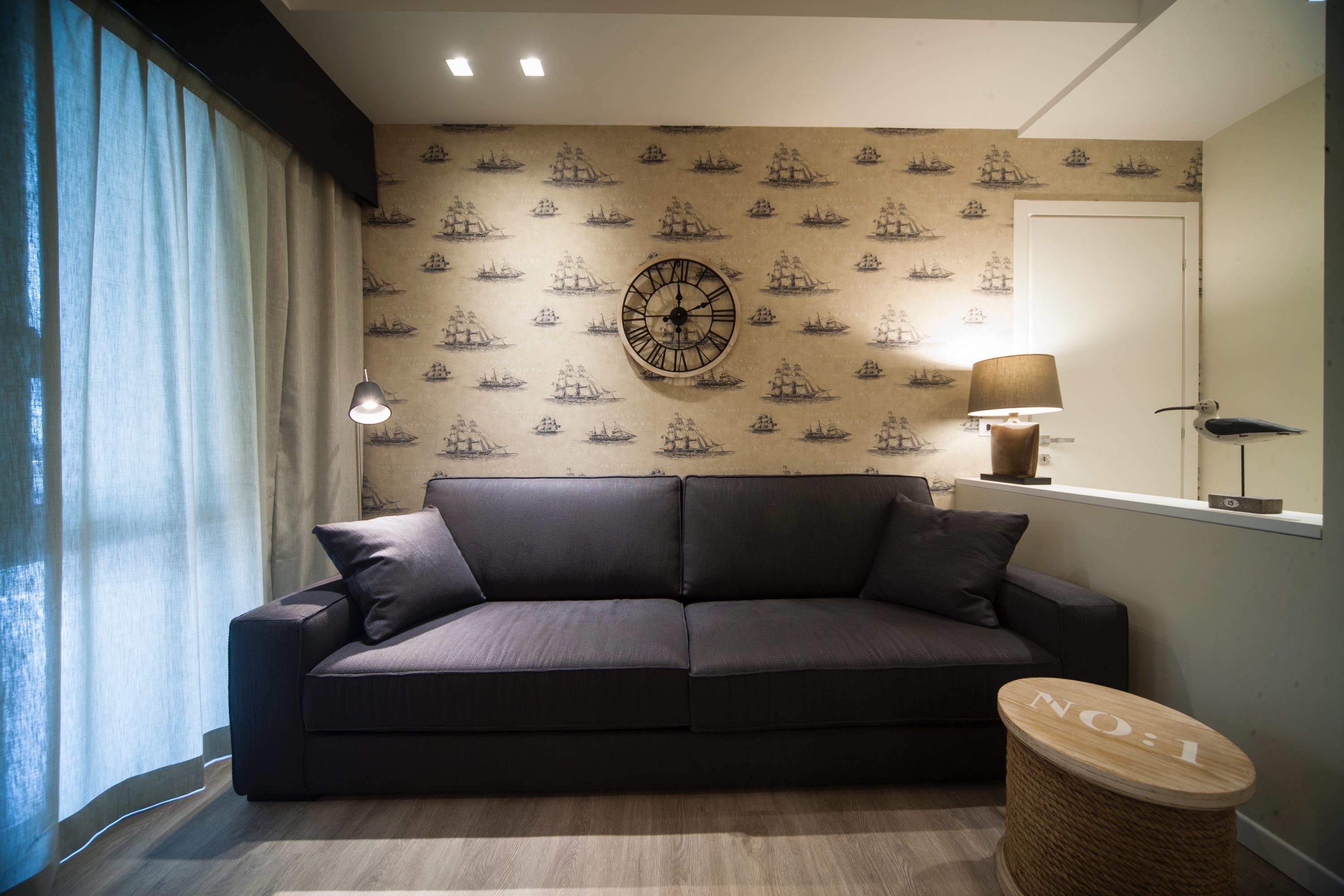 A modern living room with a dark grey sofa, a round wall clock, and a ship-themed wallpaper.