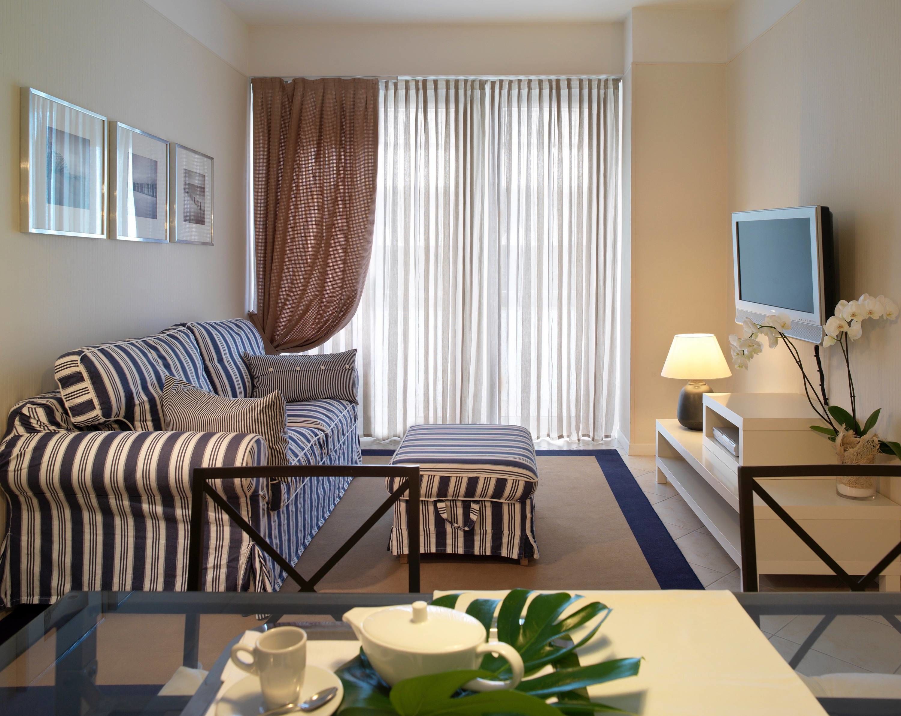 Una habitación de hotel con un sofá a rayas, un pequeño otoman, un televisor sobre un mueble bajo, una mesa de centro con tetera y taza, y una ventana con cortinas.
