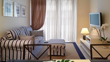 Una habitación de hotel con un sofá a rayas, un pequeño otoman, un televisor sobre un mueble bajo, una mesa de centro con tetera y taza, y una ventana con cortinas.