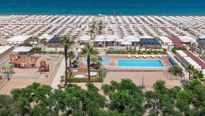 Un gran complejo turístico con numerosas sombrillas en la playa, una piscina central y una zona de terraza de madera.
