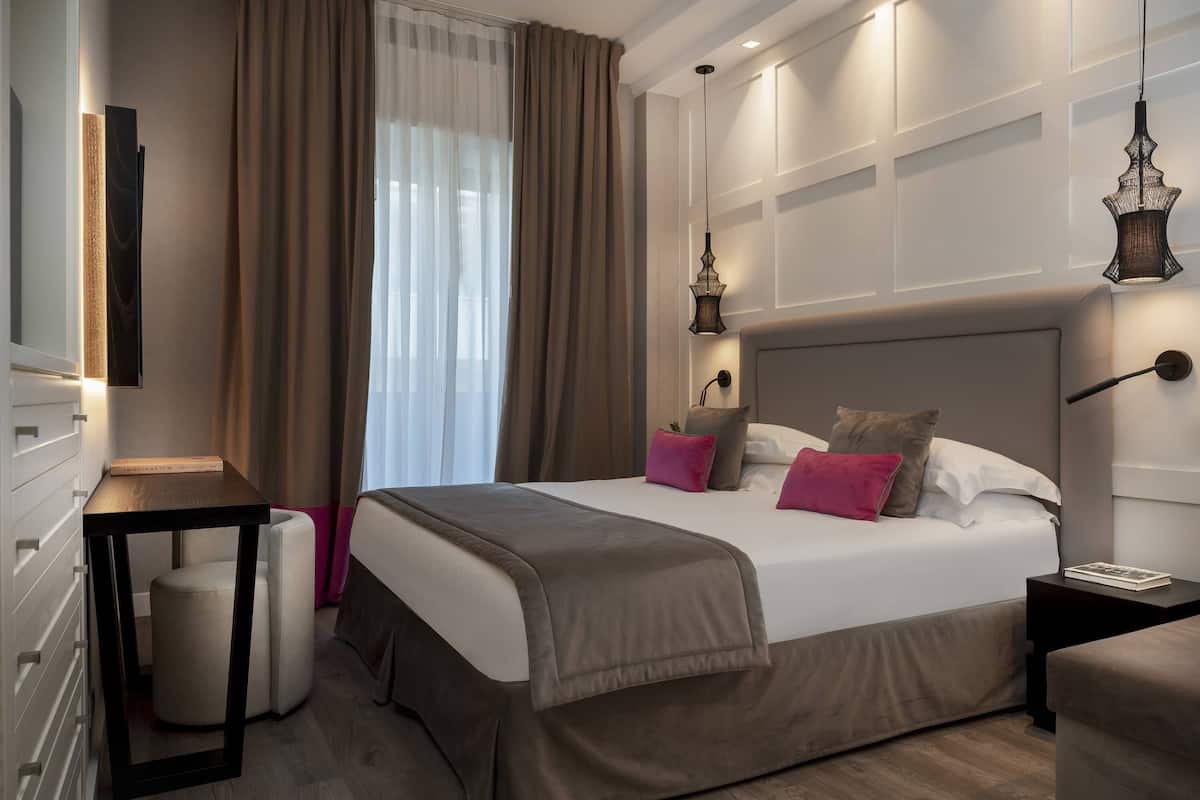 A modern hotel room with a large bed, bedside tables, a bedside lamp, and a view of the outdoors through sheer curtains.