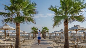 Un paseo marítimo bordeado de palmeras y sombrillas de playa, que conduce a una zona de playa con tumbonas.
