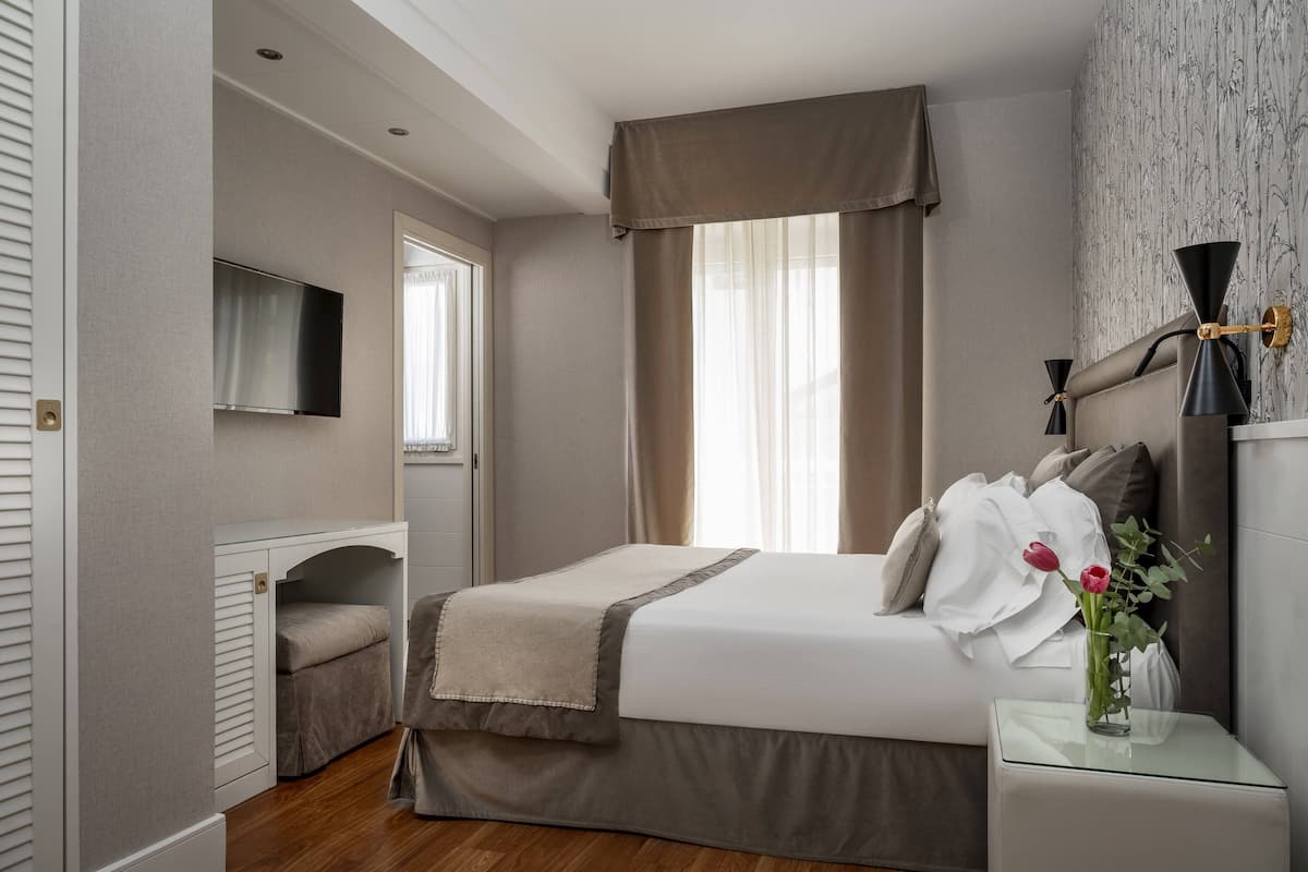 Une chambre à coucher avec un lit, un téléviseur fixé au mur, une fenêtre avec des rideaux et une table de chevet ornée d’un vase de fleurs.
