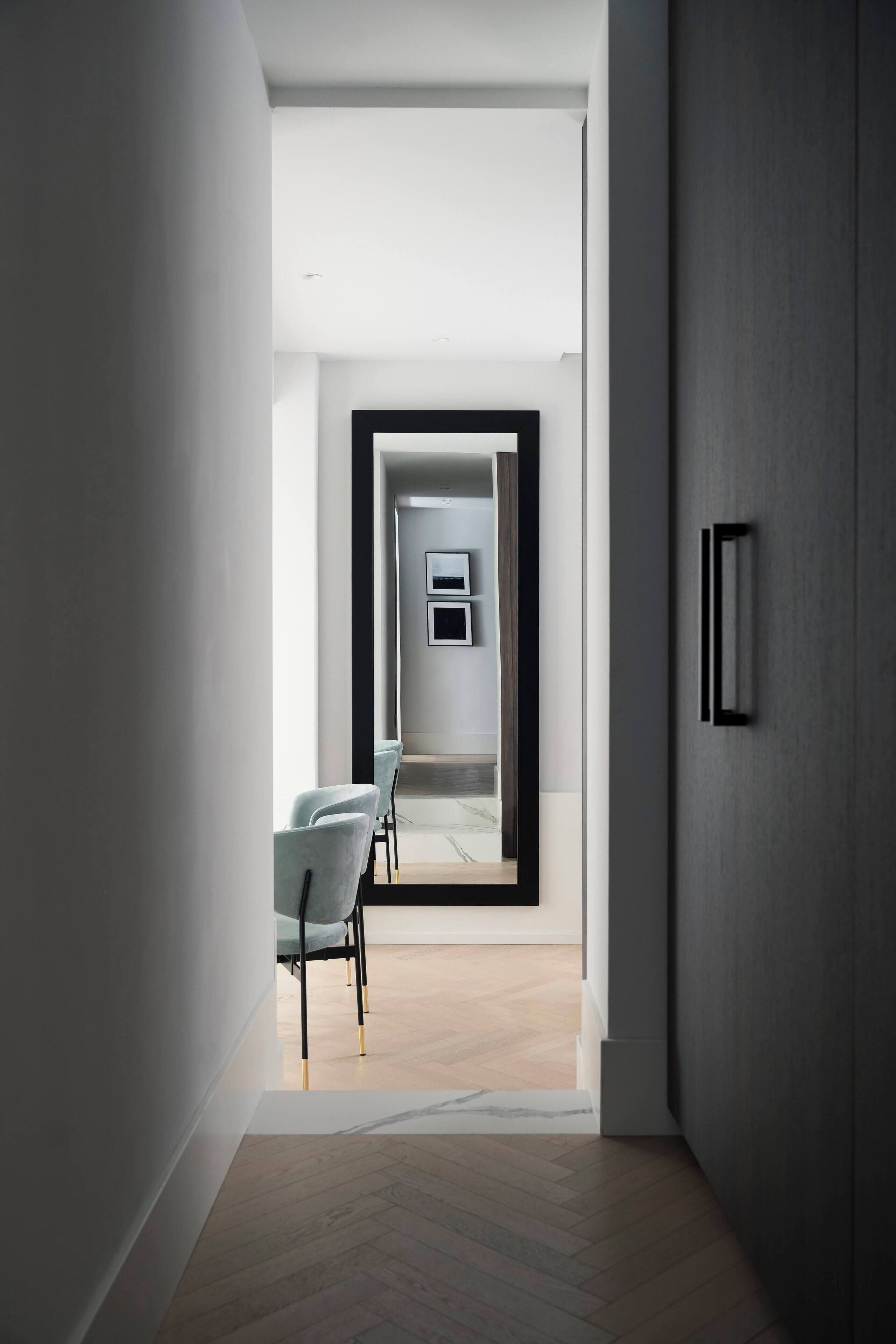Un couloir moderne avec un plancher en bois, un mur gris et une porte donnant sur une pièce où se trouvent une chaise blanche et un téléviseur à écran plat fixé au mur.