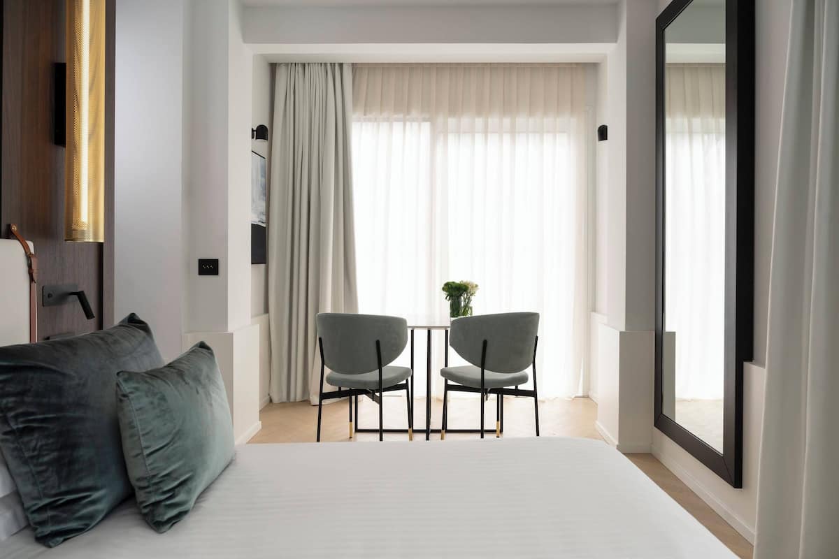 Une chambre d’hôtel avec un lit, deux chaises, une table, un grand miroir et une vue sur l’extérieur à travers des rideaux transparents.