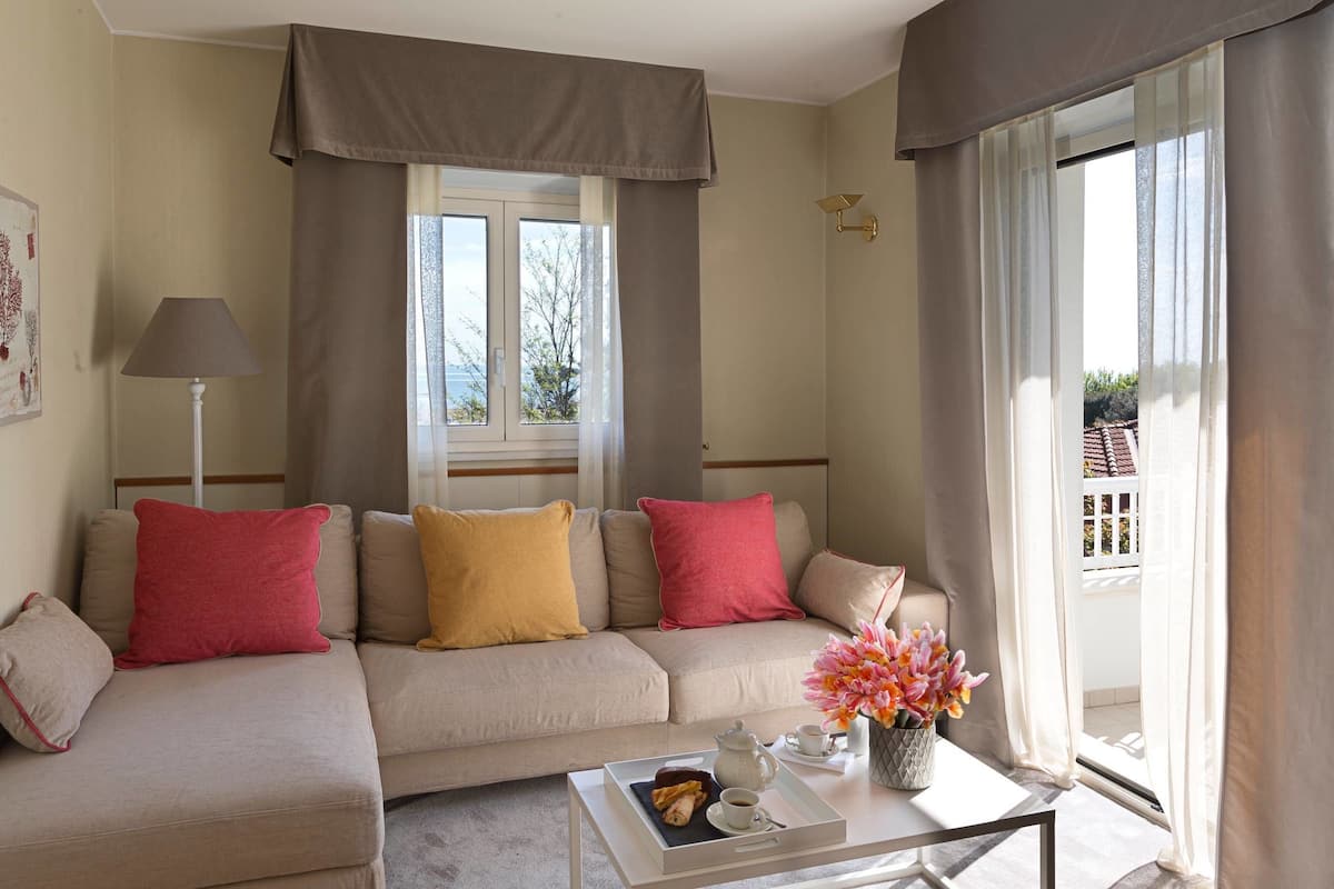 Un salon avec un canapé beige, des coussins rouges et jaunes, une table basse blanche sur laquelle se trouve un plateau de pâtisseries, un vase de fleurs et une lampe.