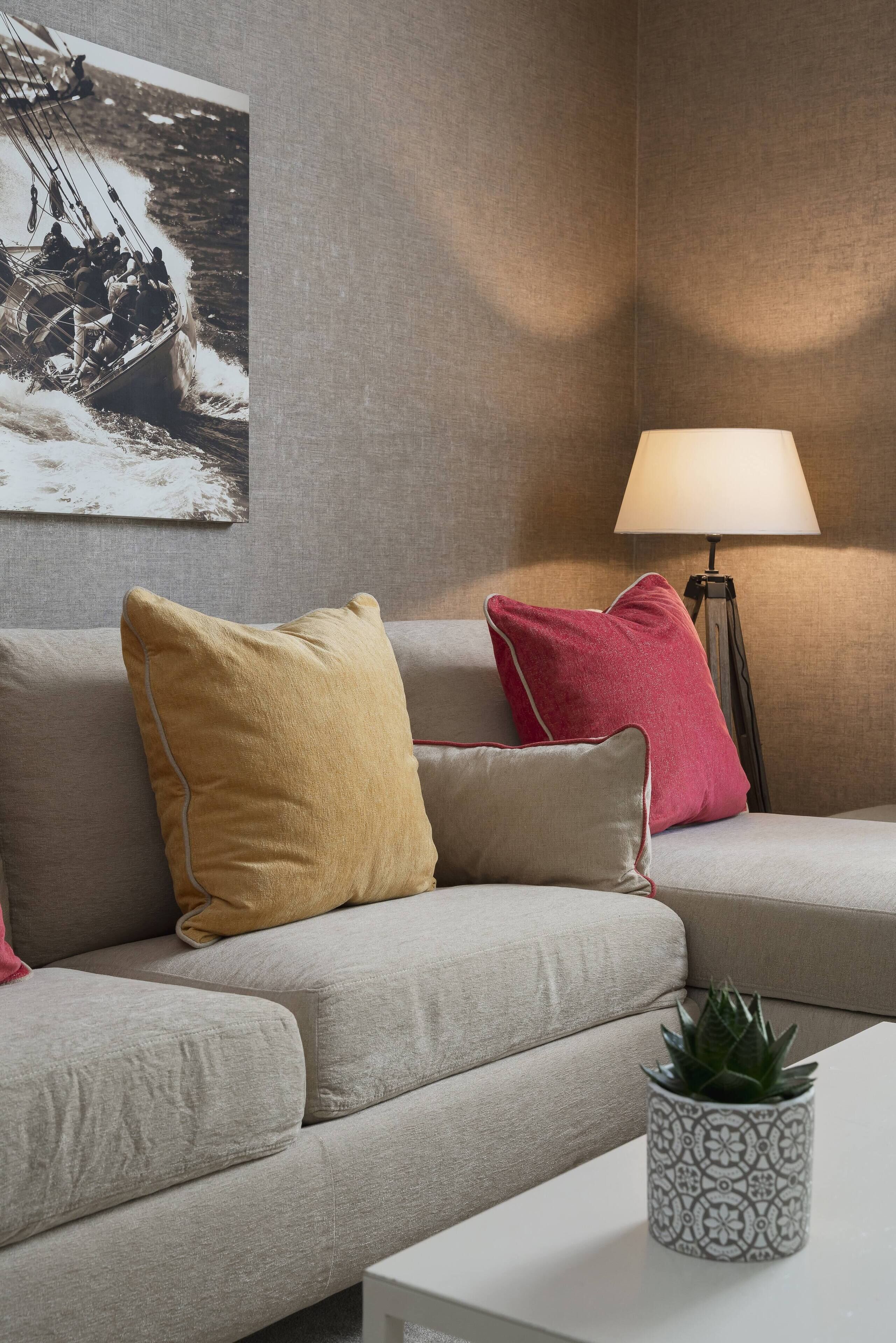 En moderne stue med en grå sofa, en rød og gul pude, en gulvlampe og en lille potteplante.